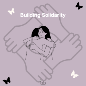 Title - Building Solidarity Image - a person hugging another, with a faded image in the background of 4 hands joined together.